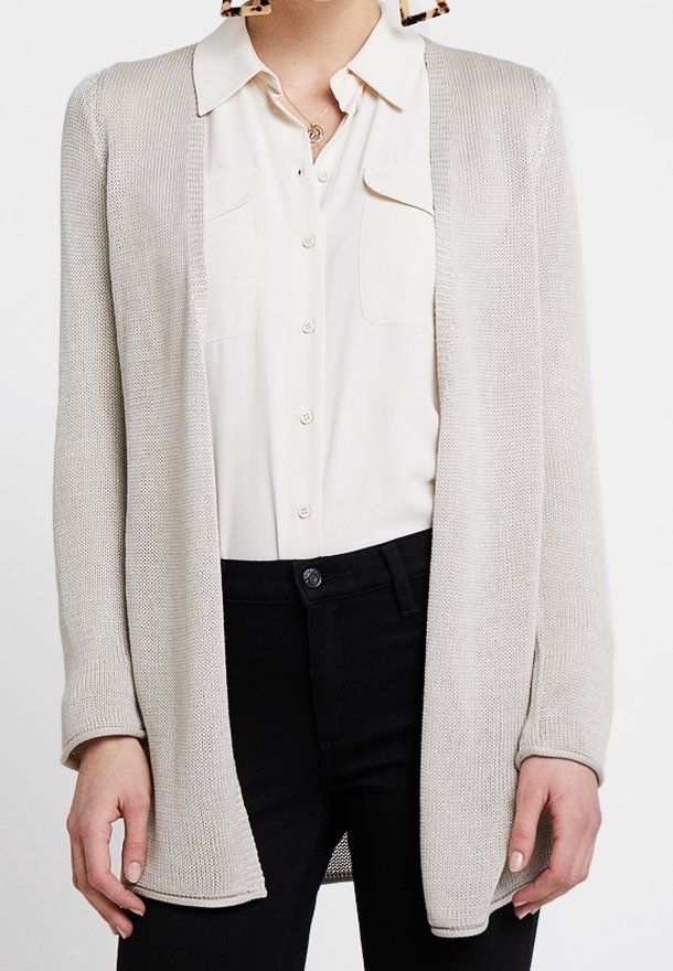 Cardigan aperto di un beige chiaro realizzato in tessuto lavorato a maglia, con maniche lunghe e vestibilità rilassata, indossato sopra una camicia button-up color crema e pantaloni scuri.