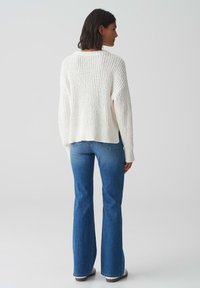 Witte gebreide trui met een losse pasvorm en ribbelstructuur, gecombineerd met uitlopende blauwe denim jeans. De achterkant toont details van de outfit.