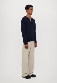 Námorník pletený pulovr s límečkem, spárovaný s širokými béžovými kalhotami. Tento outfit se vyznačuje volným střihem a texturovanou tkaninou. Černé boty dokreslují celý vzhled.