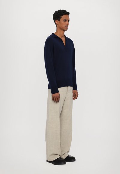Pullover blu navy a maglia con colletto, abbinato a pantaloni beige larghi. L'outfit presenta una vestibilità rilassata e un tessuto testurizzato. Scarpe nere completano il look.