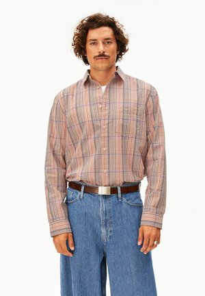 Mann mit lockigem Haar und Schnurrbart, der ein beige kariertes Hemd mit Knöpfen trägt, das in blaue Jeans mit braunem Gürtel gesteckt ist, vor weißem Hintergrund.