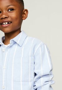 Camicia azzurra chiara con righe verticali bianche. Tessuto di cotone, colletto, taschino frontale e dettagli di chiusura con bottoni. La texture appare morbida.