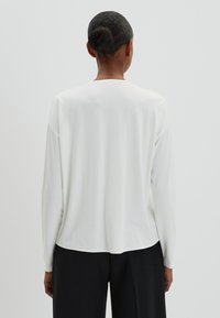 Maglietta bianca a maniche lunghe con collo rotondo, texture morbida, vestibilità rilassata e orlo posteriore leggermente più lungo, indossata con pantaloni neri.