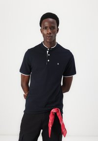 Marine polo shirt met korte mouwen, witte kraagafwerking en een drieknoopsluiting. Bevat een klein logo en een rode bandana die om de taille is geknoopt.