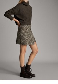 Maglione a collo alto marrone, mini gonna a quadri con bottoni e stivali alla caviglia marroni con lacci. I materiali includono maglia e tessuto intrecciato.