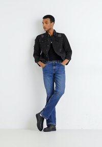 Jachetă din denim negru cu patch-uri, tricou negru, blugi drepți albaștri și teniși negri, stând pe un fundal alb.