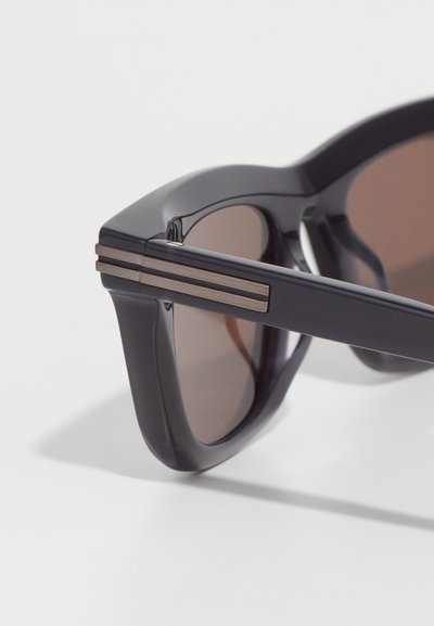 Sunglasses with a black plastic frame, brown lenses, and silver accent stripes on the temples. Geometric shape with smooth texture.
