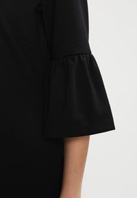 Svart topp med en klockformad ärm, gjord av mjukt tyg. Ärmen har en sammandragning vilket ger textur och volym.