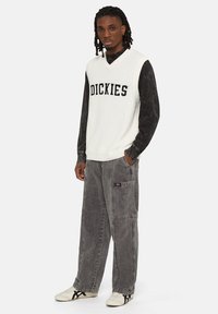 Vest tricotat alb cu inscripția "DICKIES" în litere negre, purtat peste o cămașă cu mânecă lungă neagră. Pantaloni cargo gri, largi, și sneakers albi.