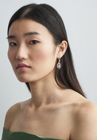 Uhani v zlati barvi z visečim dizajnom, z ovalnim belim kamnom, obkroženim z izvrstnimi detajli, ki jih nosi model z ravnimi črnimi lasmi.