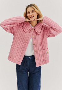 Růžová prošívaná bunda s volánovým límcem, texturovanými vertikálními pruhy, dřevěnými knoflíky a předními kapsami, doplněná bílým topem a modrými džíny.