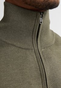 Groene gebreide zip-up kledingstuk met een hoge kraag, ribbeltextuur bij de kraag en rits, en een gladde lichaamsstructuur.