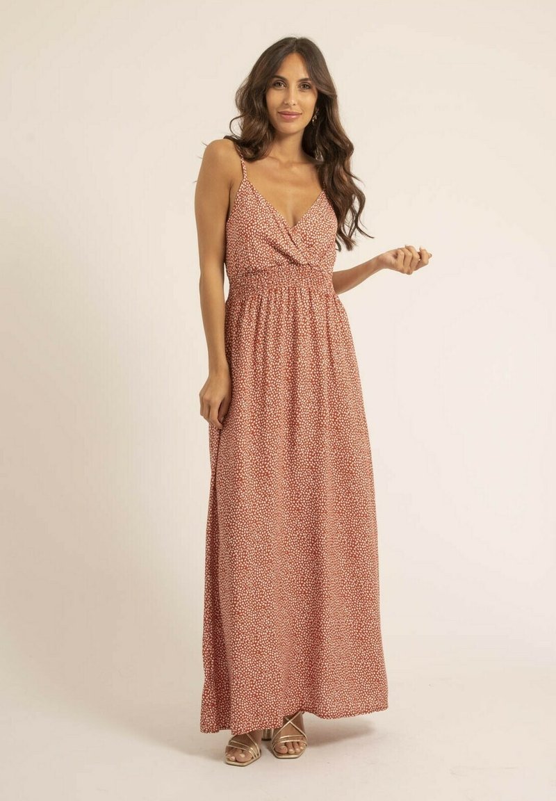 Robe maxi en rouille avec un motif de pois blancs. Elle présente un corsage torsadé, des bretelles réglables et une jupe fluide. Portée avec des sandales à lanières.