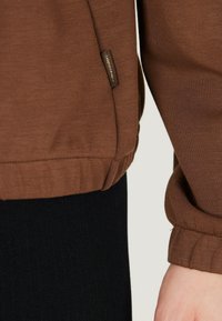 Sweatshirt marron avec une finition texturée, poignets côtelés et une étiquette logo discrète sur le côté, associé à un pantalon noir à côtes.