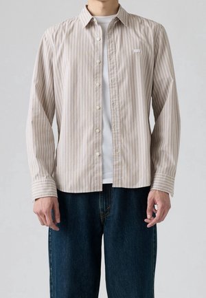 Homme portant une chemise beige et blanche à rayures verticales, boutonnée, sur un t-shirt blanc et un jean bleu foncé, sur fond gris.