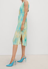 Ein ärmelloses, knielanges Kleid mit einem blau-gelben Aquarellmuster, strukturiertem Stoff und einem Seitenschlitz. Kombiniert mit himmelblauen Absätzen.