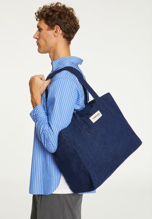 Sac fourre-tout navy en matériau feutré avec deux poignées, de forme rectangulaire et une étiquette latérale. Présente une surface texturée.