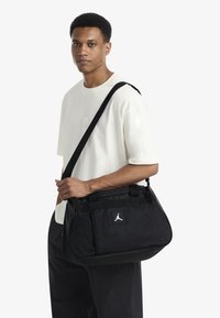 Black fabric duffel bag with structured shape, top handles, and adjustable shoulder strap. Features a prominent white logo on the front.