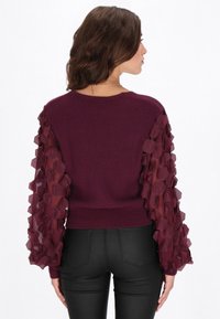 Viininpunainen neulepaita, jossa on ribbineulosta ja pussimaiset läpikuultavat hihat, joissa on kerroksittain kuusikulmaisia kuvioita. Istuvamalli pyöreällä pääntiellä.