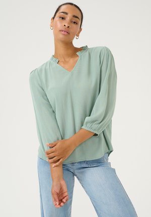 Light green, V-neck blouse with a ruffled collar and 3/4 puff sleeves. Textured fabric, relaxed fit, paired with light blue jeans.