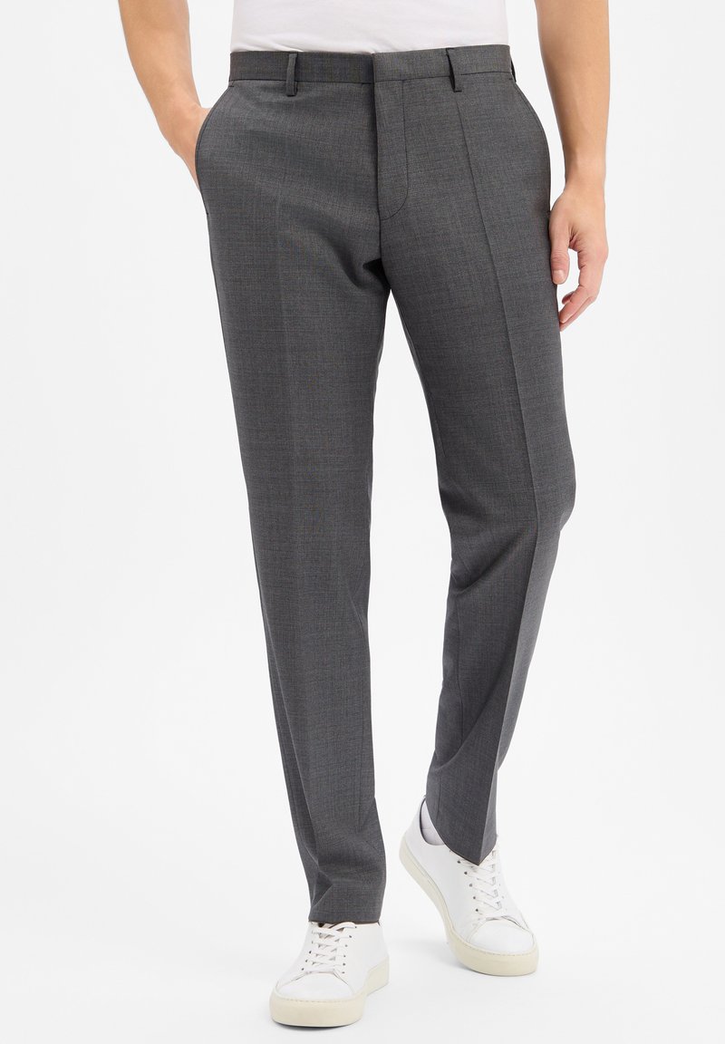 Man wearing gray tailored trousers with front crease and white sneakers, standing with hands in pockets on white background.