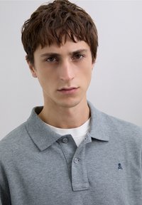 Polo gris en tissu texturé, avec un col et deux boutons. Un petit logo bleu marine représentant une tête de mort avec des os croisés se trouve sur le côté gauche de la poitrine.