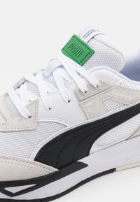 Basket de sport blanche et grise en matières mesh et suède, avec des accents noirs, des lacets blancs plats, et une étiquette logo verte sur la languette.