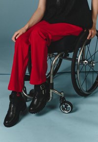 Black ankle boots with thick soles and two gold buckles, paired with red pants, seated in a wheelchair on a light blue background.