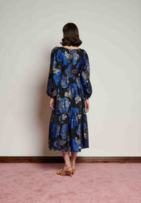 Μαύρο midi φόρεμα με λουλουδάτο μοτίβο, μπλε και χρυσές διακοσμήσεις, μανίκια φουσκωτά, φούστα με στρώσεις και κλείσιμο με φερμουάρ στην πλάτη. Φορεμένο με καφέ πέδιλα με τακούνι.