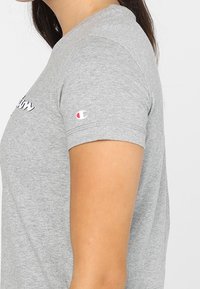 Camiseta de manga corta gris con cuello redondo, con un pequeño logo en el pecho y un logo de Champion en la manga izquierda. Material de algodón suave.