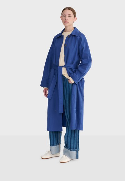 Femme debout portant un trench-coat bleu ceinturé, une chemise beige, un jean large retroussé et des baskets blanches sur un fond uni.
