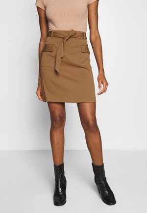 Brauner Baumwollrock mit Taillenband, zwei Vordertaschen und figurbetontem Schnitt, kombiniert mit schwarzen Lack-Stiefeletten.