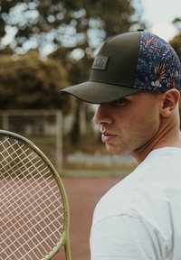Black cap with a floral pattern on the side, paired with a white T-shirt. A green tennis racket is held beside the person.