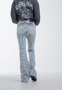 Jeans acampanados en azul claro con un patrón de rayas de cebra, que presentan un logo bordado de "Ed Hardy" en la cinturilla y en los bolsillos traseros.