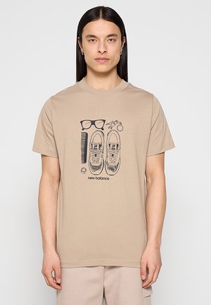Mann mit langen schwarzen Haaren trägt ein beiges New Balance T-Shirt mit Illustrationen von Sneakers, Brille, Kamm, Schlüsseln und Ring.