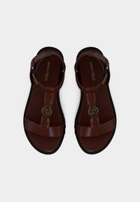 Καφέ συνθετικά επίπεδα σανδάλια με μαύρες σόλες, που διαθέτουν ρυθμιζόμενους ιμάντες στον αστράγαλο και διακοσμητικό χρυσό στοιχείο στο κέντρο.