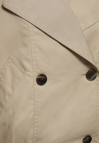 Detaliu al paltonului trench bej care arată nasturii dubli, reverul larg și cusăturile buzunarului frontal în unghi.