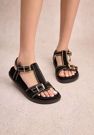 Pieds portant des sandales noires avec des clous dorés et des boucles sur un sol clair texturé, montrant des ongles de pieds vernis.