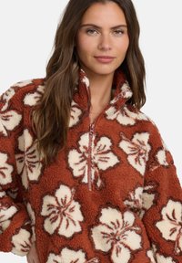 Pullover in pile ruggine con motivo floreale crema. Presenta un collo a mezze zip e una superficie morbida e testurizzata. Accentuato da contorni scuri.