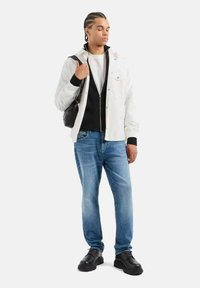 Giacca leggera bianca sopra un felpa con zip nera, abbinata a jeans blu e scarpe chunky nere. Tiene una piccola borsa nera.