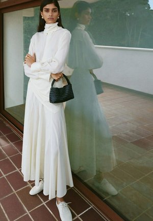 Femme portant une longue robe blanche et des chaussures blanches, tenant un sac à main noir tressé, debout à côté d'un mur de verre avec son reflet.