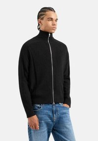 Maglione nero con collo alto e zip, con texture a costine, dotato di una zip argentata e polsini aderenti, abbinato a jeans in denim blu chiaro.