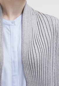 Jasnoszary kardigan w prążki, wykonany z miękkiego materiału, z kołnierzem szalowym i gładką niebieską koszulą pod spodem, z widocznym zapięciem na guziki.