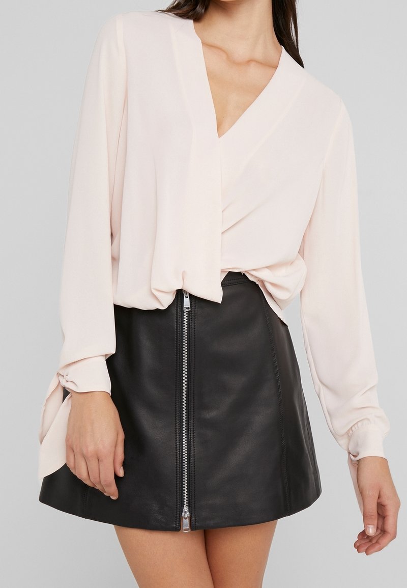 Femme portant un chemisier rose pâle à manches longues, rentré dans une mini-jupe en cuir noire avec un zip sur le devant.