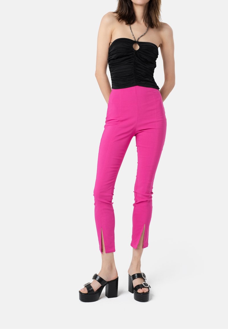 Femme portant un haut noir à bretelles froncé, un pantalon slim rose vif à fentes sur le devant des jambes, et des sandales noires à talons compensés avec boucles.
