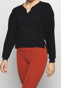 Schwarzes, kurz geschnittenes Langarmtop mit V-Ausschnitt und strukturiertem Rippmaterial. Mit seitlichen Schlitzen und eng anliegendem Bündchen versehen. Getragen mit roten Leggings.