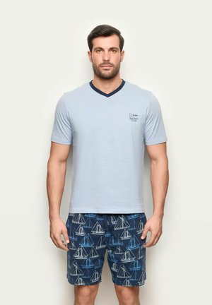 Mann trägt hellblaues V-Neck T-Shirt mit "Racing Club"-Logo und marineblaue Shorts mit weiß-blauem Segelbootmuster, steht vor einfarbigem Hintergrund.