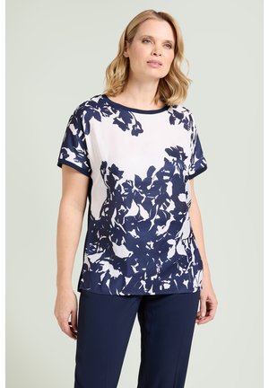 Kvinde iført en kortærmet marineblå og hvid bluse med blomsterprint kombineret med marineblå bukser, stående foran en ensartet lys baggrund.