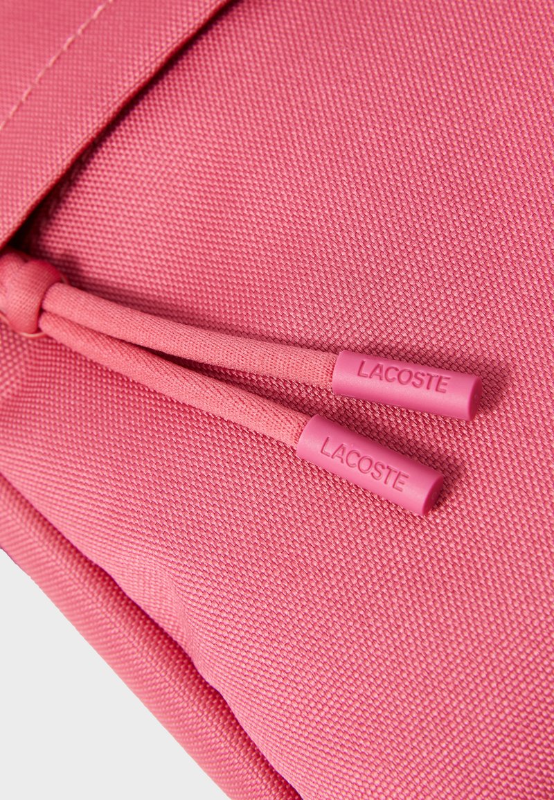 Détail d'un sac à dos rose montrant un tissu texturé avec une finition lisse et deux tirettes de fermeture éclair rose marquées "LACOSTE."