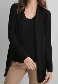 Svart, långärmad, öppen cardigan i mjukt tyg, kombinerad med en åtsittande svart topp och ljusbruna byxor. Enkel design, utan mönster.